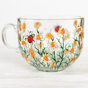 Puede incluir: Una taza de t&eacute; de vidrio transparente con asa, decorada con margaritas pintadas a mano, tallos verdes y una mariquita. Las flores tienen p&eacute;talos blancos, centros naranjas y detalles azules. Una taza encantadora.