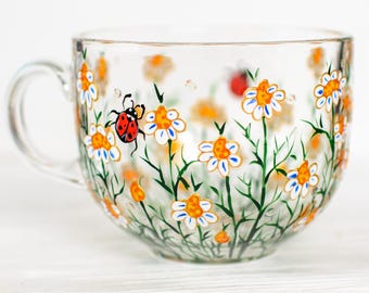 May include: A clear glass teacup featuring a hand-painted design of white daisies with yellow centers, green stems, and a single red ladybug with black spots. The cup has a curved handle and a wide, rounded bowl shape with a smooth glass finish.