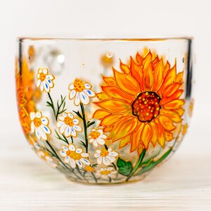 Op de afbeelding: Een doorzichtige glazen mok met een handgeschilderd ontwerp van een grote, levendige gele en oranje zonnebloem naast verschillende kleinere witte madeliefjes met gele harten en groene stelen, alles in een gedetailleerd bloemenpatroon.