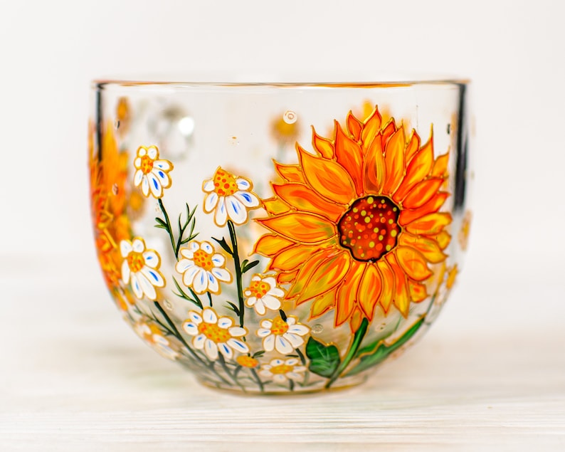 Op de afbeelding: Een doorzichtige glazen mok met een handgeschilderd ontwerp van een grote, levendige gele en oranje zonnebloem naast verschillende kleinere witte madeliefjes met gele harten en groene stelen, alles in een gedetailleerd bloemenpatroon.
