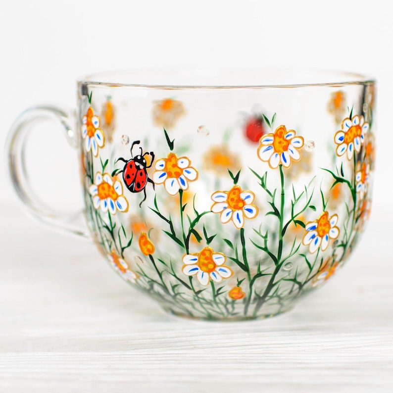Puede incluir: Una taza de t&eacute; de vidrio transparente con asa, decorada con margaritas blancas pintadas a mano con centros naranjas y detalles azules, tallos verdes y una mariquita roja. La taza est&aacute; dise&ntilde;ada para t&eacute; o sopa.