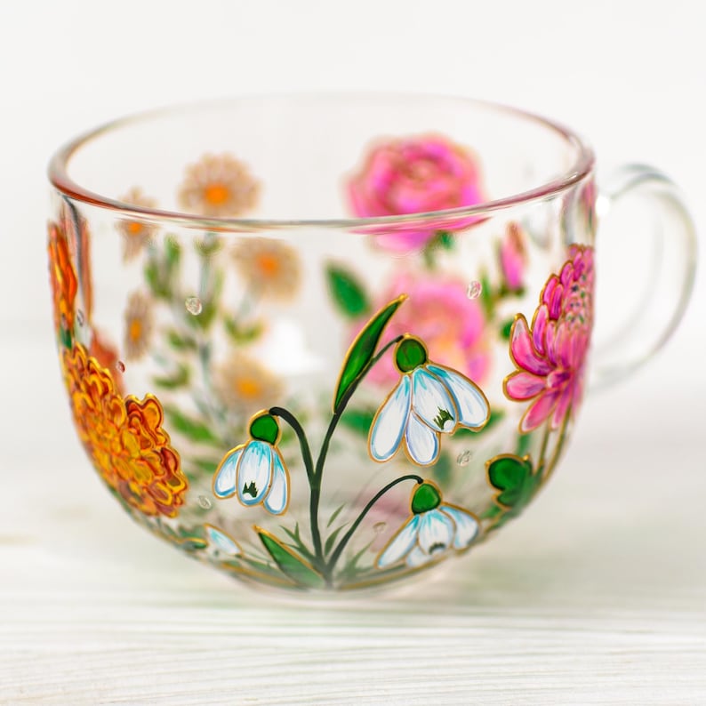 K&ouml;nnte beinhalten: Eine transparente Teetasse mit Henkel, handbemalt mit bunten Blumen. Das florale Design umfasst rosa Rosen, orangefarbene Ringelblumen und wei&szlig;e Schneegl&ouml;ckchen mit gr&uuml;nen Bl&auml;ttern. Die Tasse steht auf einer wei&szlig;en Oberfl&auml;che.