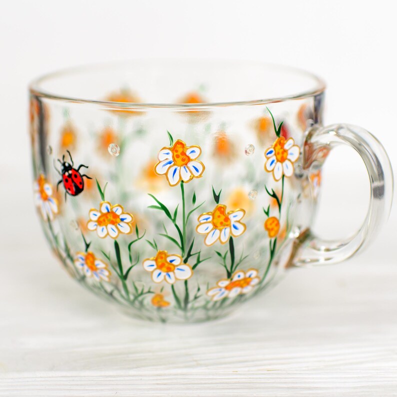 Puede incluir: Una taza de t&eacute; de vidrio transparente con asa, pintada a mano con margaritas blancas y naranjas y tallos verdes. Una mariquita roja y negra tambi&eacute;n est&aacute; pintada en el lateral de la taza. La taza est&aacute; sobre una superficie blanca.