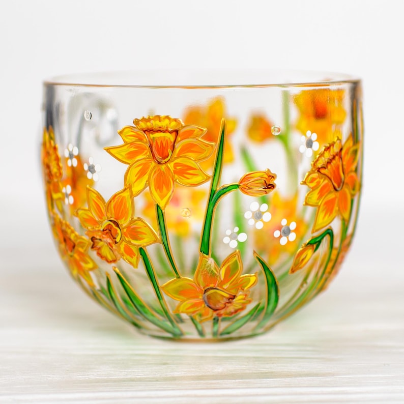 May include: Clear glass bowl decorated with hand-painted yellow daffodils and green stems. The floral design features orange and yellow petals with white accents. A decorative piece for the home.