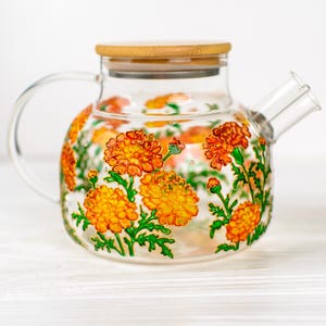 May include: A clear glass teapot featuring a hand-painted design of vibrant orange marigold flowers with green stems and leaves. The teapot includes a circular bamboo lid and a curved glass handle, showcasing a transparent body and spout.
