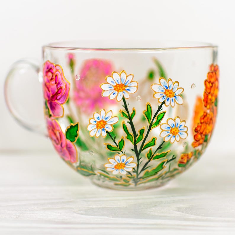 K&ouml;nnte beinhalten: Eine transparente Teetasse mit Henkel, handbemalt mit farbenfrohen Blumenmustern. Die Tasse zeigt rosa Rosen, wei&szlig;e G&auml;nsebl&uuml;mchen mit blauen Zentren und orangefarbene Bl&uuml;ten. Gr&uuml;ne Bl&auml;tter und goldene Akzente vervollst&auml;ndigen das Design.