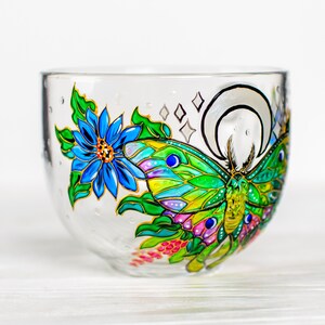 May include: A clear glass bowl with a hand-painted design featuring a blue flower, a green and yellow butterfly, and a crescent moon. The bowl has a rounded shape and is decorated with colorful details and gold accents.