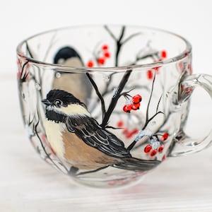 May include: A clear glass mug featuring a hand-painted design of a black-capped chickadee perched on a branch. The artwork includes black tree branches, small clusters of bright red berries, and white accents representing snow on the branches.