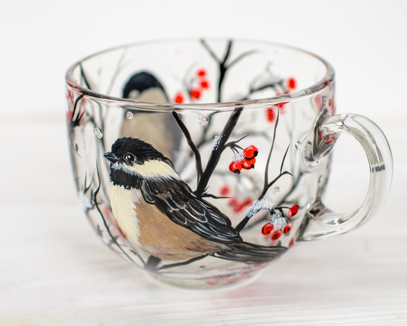 May include: A clear glass mug featuring a hand-painted design of a black-capped chickadee perched on a branch. The artwork includes black tree branches, small clusters of bright red berries, and white accents representing snow on the branches.
