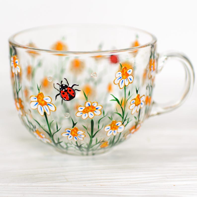 Puede incluir: Una taza de t&eacute; de vidrio transparente con asa, pintada a mano con un dise&ntilde;o floral. La taza presenta margaritas blancas con centros naranjas y tallos verdes, junto con una mariquita roja y negra. La taza est&aacute; sobre una superficie blanca.