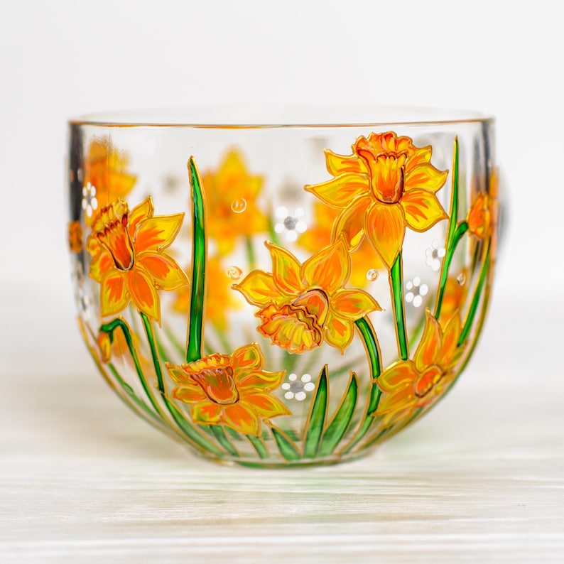 May include: A clear glass teacup with a hand-painted design of yellow daffodils with orange centers and green stems. The flowers are arranged around the cup, with small white accents. The cup has a handle and is set against a white background.