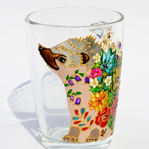 May include: A glass mug with a hand-painted design of a hedgehog with colorful flowers. The hedgehog is pink with a gray face and brown nose. The flowers are red, orange, yellow, green, blue, and purple.