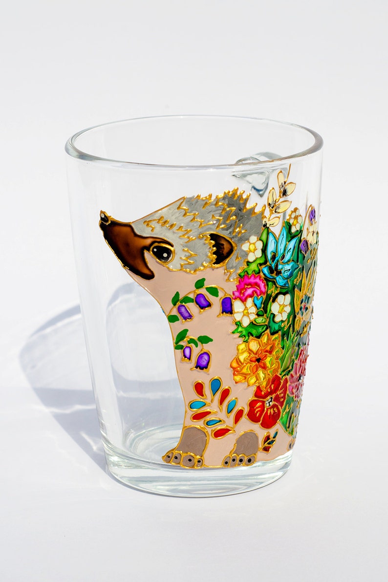 May include: A glass mug with a hand-painted design of a hedgehog with colorful flowers. The hedgehog is pink with a gray face and brown nose. The flowers are red, orange, yellow, green, blue, and purple.