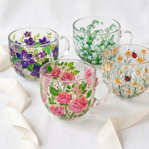 Set of four hand-painted birth flower glass mugs with rose, violet, lily of the valley, and daisy designs, styled as matching bridesmaid gifts with an ivory ribbon.