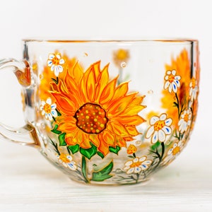Op de afbeelding: Een doorzichtige glazen mok met een handgeschilderd ontwerp van levendige gele en oranje zonnebloemen met groene bladeren en kleine witte bloemetjes. Het kunstwerk loopt rond de buitenkant van de transparante beker met een gebogen oor.