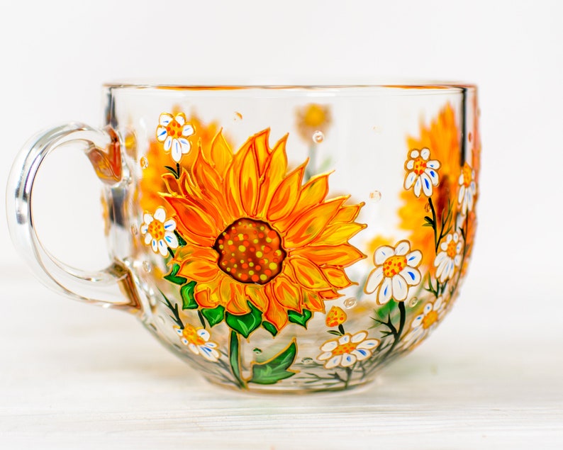 Op de afbeelding: Een doorzichtige glazen mok met een handgeschilderd ontwerp van levendige gele en oranje zonnebloemen met groene bladeren en kleine witte bloemetjes. Het kunstwerk loopt rond de buitenkant van de transparante beker met een gebogen oor.