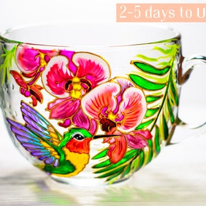 May include: A clear glass teacup with a large handle, hand-painted with vibrant pink orchids, green leaves, and a colorful hummingbird. The design is accented with gold outlines, creating a decorative and artistic look.