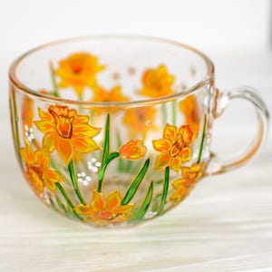 May include: A clear glass teacup with a handle, adorned with hand-painted yellow and orange daffodils and green stems. The floral design wraps around the cup, creating a cheerful and artistic look. The cup is perfect for serving tea or coffee.