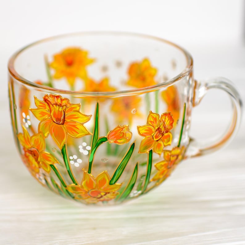 May include: A clear glass teacup with a handle, adorned with hand-painted yellow and orange daffodils and green stems. The floral design wraps around the cup, creating a cheerful and artistic look. The cup is perfect for serving tea or coffee.