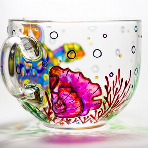 May include: A clear glass teacup with a painted underwater scene. The design includes a colorful fish, a pink seashell, red coral, and black bubbles. The handle is clear and curved.