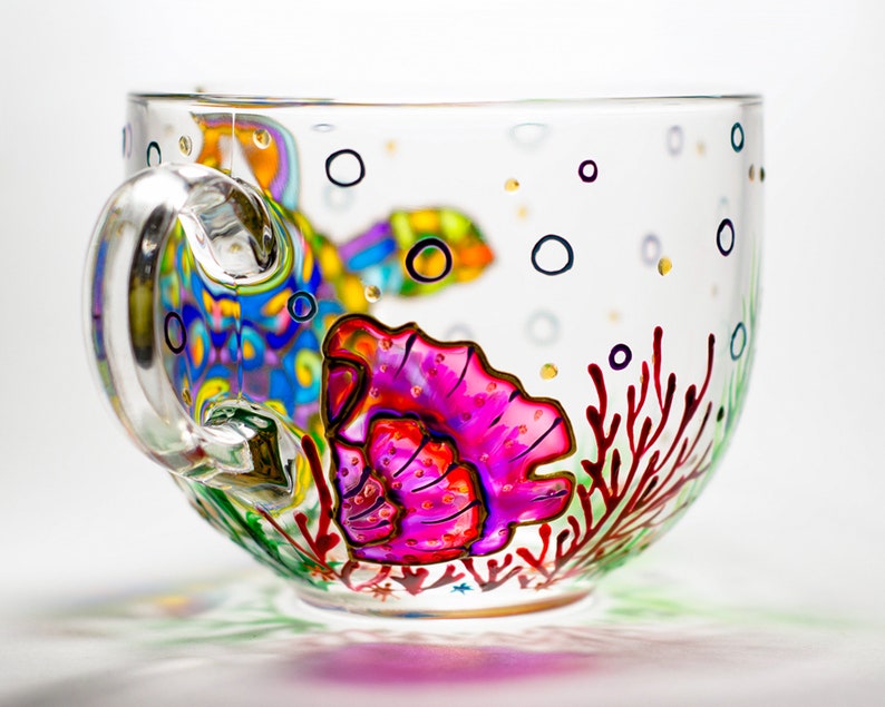 May include: A clear glass teacup with a painted underwater scene. The design includes a colorful fish, a pink seashell, red coral, and black bubbles. The handle is clear and curved.