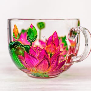 Puede incluir: Una taza de t&eacute; de vidrio transparente con un asa grande, pintada a mano con un vibrante dise&ntilde;o de flor de loto. El loto presenta tonos de rosa, verde y naranja, con detalles dorados. La taza est&aacute; sobre un fondo blanco.