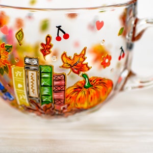Puede incluir: Una taza de cristal transparente con dise&ntilde;os pintados a mano que incluyen una calabaza naranja brillante, una pila de libros coloridos, un girasol, hojas de oto&ntilde;o, peque&ntilde;as cerezas rojas y diminutos corazones rosas en la superficie.
