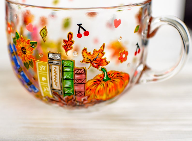 Puede incluir: Una taza de cristal transparente con dise&ntilde;os pintados a mano que incluyen una calabaza naranja brillante, una pila de libros coloridos, un girasol, hojas de oto&ntilde;o, peque&ntilde;as cerezas rojas y diminutos corazones rosas en la superficie.