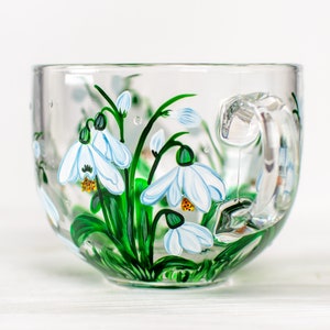 May include: A clear glass mug featuring hand-painted white snowdrop flowers with green stems and leaves. The floral design wraps around the exterior of the cup, showcasing delicate white petals with yellow centers and vibrant green foliage.
