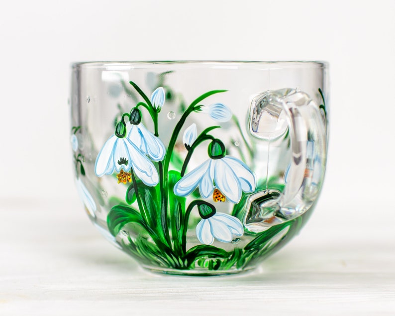 May include: A clear glass mug featuring hand-painted white snowdrop flowers with green stems and leaves. The floral design wraps around the exterior of the cup, showcasing delicate white petals with yellow centers and vibrant green foliage.