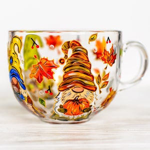 Könnte beinhalten: Eine transparente Glas-Tasse mit Henkel, verziert mit bunten Herbst-Zwergen und Herbstblättern. Die Zwerge tragen Hüte in Braun-, Orange- und Gelbtönen. Ein Kürbis wird von einem der Zwerge gehalten. Ideal für ein warmes Getränk.
