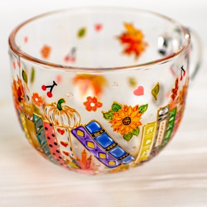 Puede incluir: Una taza de cristal transparente con un dise&ntilde;o pintado a mano de libros coloridos, una calabaza blanca y un girasol amarillo brillante. El exterior est&aacute; decorado con peque&ntilde;as cerezas rojas, hojas de oto&ntilde;o y corazones rosas dispersos.