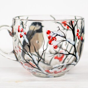 May include: A clear glass mug featuring a hand-painted winter scene with a small bird perched on black branches. The design includes clusters of bright red berries and white snow accents painted on the exterior surface of the transparent glass.