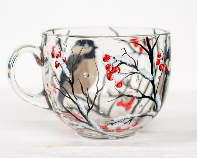 May include: A clear glass mug featuring a hand-painted winter scene with a small bird perched on black branches. The design includes clusters of bright red berries and white snow accents painted on the exterior surface of the transparent glass.