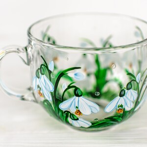 May include: A clear glass teacup featuring a hand-painted design of white snowdrop flowers with green stems and leaves. The floral pattern wraps around the exterior of the cup, which includes a curved handle on the left side and a wide rim.