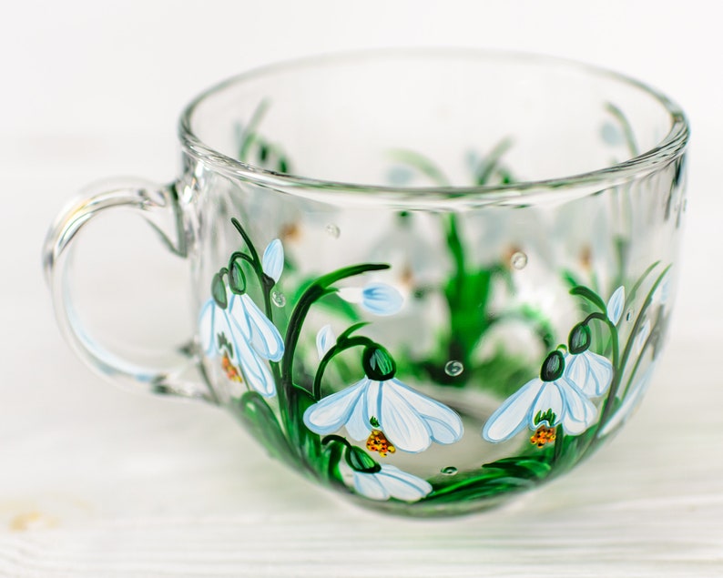 May include: A clear glass teacup featuring a hand-painted design of white snowdrop flowers with green stems and leaves. The floral pattern wraps around the exterior of the cup, which includes a curved handle on the left side and a wide rim.