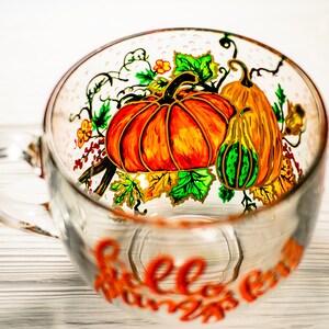 May include: A clear glass mug featuring a hand-painted design of orange pumpkins, a green and yellow gourd, and green leaves. The text "Hello Pumpkin" is written in orange script on the lower front of the mug, which has a single handle.