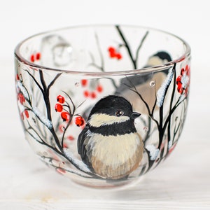 May include: A clear glass teacup featuring a hand-painted design of a black-capped chickadee perched on a snowy branch with bright red berries. The bird has a black head, white cheeks, and a tan chest, surrounded by black branches and white snow.