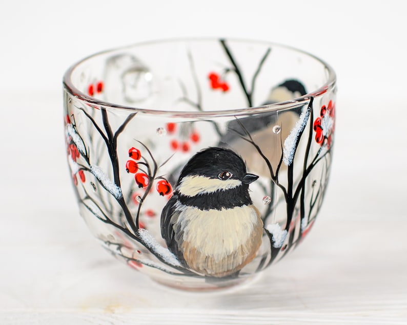 May include: A clear glass teacup featuring a hand-painted design of a black-capped chickadee perched on a snowy branch with bright red berries. The bird has a black head, white cheeks, and a tan chest, surrounded by black branches and white snow.