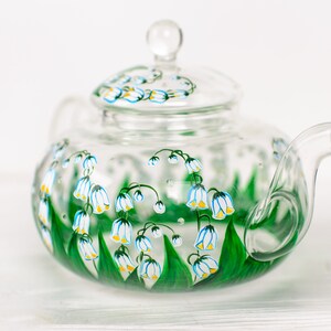 May include: A clear glass teapot with a lid, hand-painted with white and yellow lily of the valley flowers and green leaves.