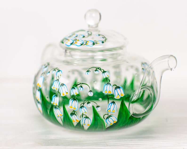 May include: A clear glass teapot with a lid, hand-painted with white and yellow lily of the valley flowers and green leaves.