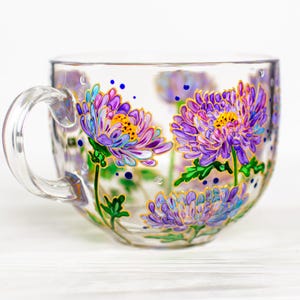 Puede incluir: Una taza de t&eacute; de vidrio transparente con asa, adornada con flores de crisantemo pintadas a mano en morado y amarillo. El dise&ntilde;o floral incluye hojas verdes y detalles dorados, creando un aspecto delicado y art&iacute;stico. La taza es ideal para servir t&eacute;.