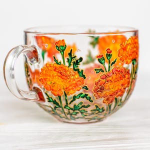 May include: A clear glass teacup with a large handle, decorated with orange and yellow marigold flowers and green leaves. The floral design is hand-painted with gold outlines, creating a vibrant and detailed botanical theme.