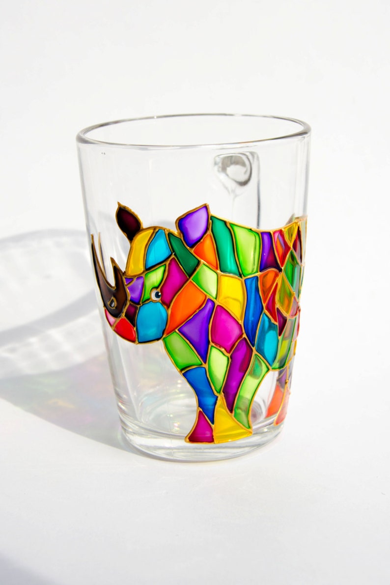 May include: A clear glass mug with a colorful rhino design. The rhino is depicted in a mosaic style, using vibrant shades of purple, green, orange, and yellow. The mug has a handle and is suitable for beverages.