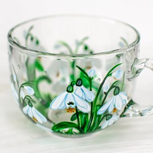 May include: A clear glass teacup featuring a hand-painted design of white snowdrop flowers with green stems and leaves. The delicate floral artwork wraps around the exterior of the cup, showcasing white petals with yellow centers and green accents.