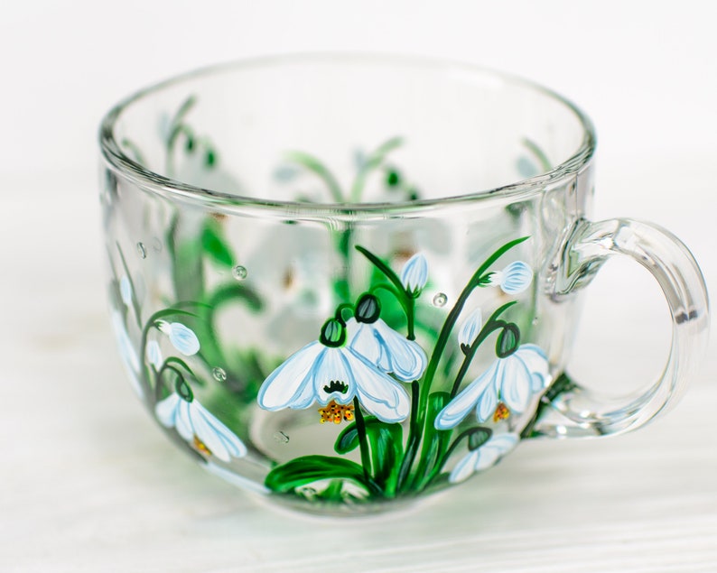 May include: A clear glass teacup featuring a hand-painted design of white snowdrop flowers with green stems and leaves. The delicate floral artwork wraps around the exterior of the cup, showcasing white petals with yellow centers and green accents.