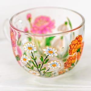 K&ouml;nnte beinhalten: Eine transparente Teetasse mit Henkel, verziert mit handgemalten Blumenmustern. Die Tasse zeigt wei&szlig;e G&auml;nsebl&uuml;mchen mit gelben Zentren, orangefarbene Ringelblumen und rosa Bl&uuml;ten. Gr&uuml;ne Bl&auml;tter und goldene Akzente vervollst&auml;ndigen das Design.