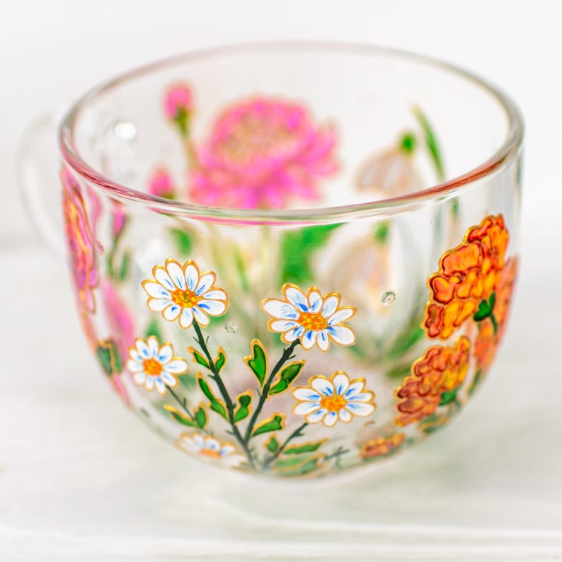 K&ouml;nnte beinhalten: Eine transparente Teetasse mit Henkel, verziert mit handgemalten Blumenmustern. Die Tasse zeigt wei&szlig;e G&auml;nsebl&uuml;mchen mit gelben Zentren, orangefarbene Ringelblumen und rosa Bl&uuml;ten. Gr&uuml;ne Bl&auml;tter und goldene Akzente vervollst&auml;ndigen das Design.