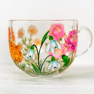 K&ouml;nnte beinhalten: Eine transparente Teetasse mit Henkel, verziert mit handgemalten Blumenmustern. Die Tasse zeigt orange, rosa und wei&szlig;e Blumen mit gr&uuml;nen Bl&auml;ttern und Stielen. Die Blumen sind auf der Au&szlig;enseite der Tasse gemalt.