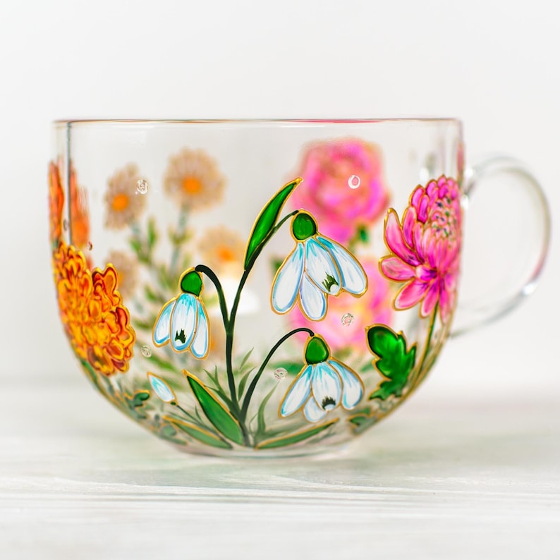 K&ouml;nnte beinhalten: Eine transparente Teetasse mit Henkel, verziert mit handgemalten Blumenmustern. Die Tasse zeigt orange, rosa und wei&szlig;e Blumen mit gr&uuml;nen Bl&auml;ttern und Stielen. Die Blumen sind auf der Au&szlig;enseite der Tasse gemalt.